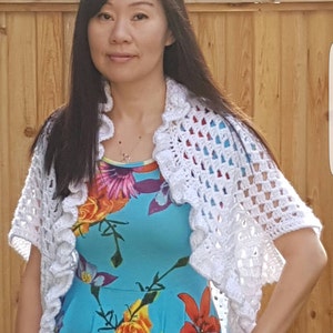 May include: A woman wearing a white crocheted shrug over a blue floral dress. The shrug has a ruffled edge and a lacy pattern.