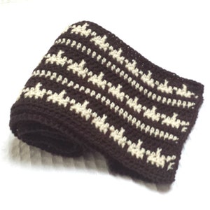 Peut inclure: Une écharpe en crochet marron et blanc avec un motif géométrique. L'écharpe est enroulée et le motif est visible sur l'extérieur du rouleau.
