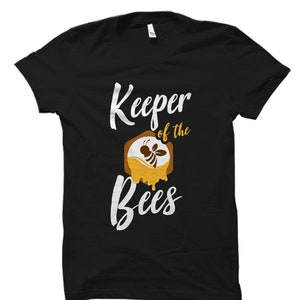 May include: Black t-shirt with white text that reads "Keeper of the Bees". A stylized image of a bee in a honeycomb is above the text.