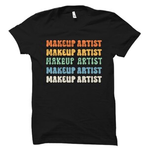 以下が含まれることがあります： 黒いTシャツ。レトロなフォントで「MAKEUP ARTIST」の文字が5回繰り返されています。テキストはオレンジ、黄色、緑、青、オフホワイトのグラデーションです。このシャツは半袖で、クルーネックです。