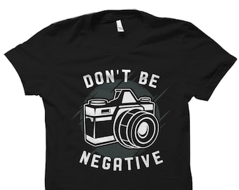 May include: Black t-shirt with a white graphic of a vintage camera and the words "DON'T BE NEGATIVE". The camera design is set against a dark gray circular background with radial lines.