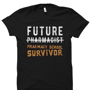 May include: Black t-shirt with white text that reads "Future Pharmacist" with a line through "Pharmacist" and the text "Pharmacy School Survivor" in orange below.