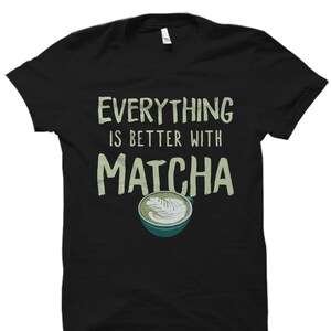 Puede incluir: Camiseta negra con el texto "Everything is better with Matcha" en verde y un latte matcha verde en una taza blanca con un platillo verde.