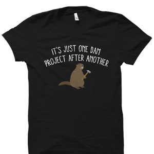 Könnte beinhalten: Schwarzes T-Shirt mit einem Cartoon-Bild eines Bibers, der einen Hammer hält, und dem Text "It's just one dam project after another."