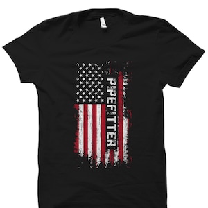 Puede incluir: Camiseta negra con un diseño de bandera estadounidense desgastada y la palabra "Pipefitter" impresa verticalmente en el lado derecho de la bandera.
