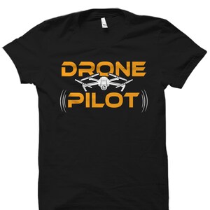Puede incluir: Camiseta negra con el texto "DRONE PILOT" en letras naranjas. Un dron blanco con una c&aacute;mara est&aacute; por encima del texto.