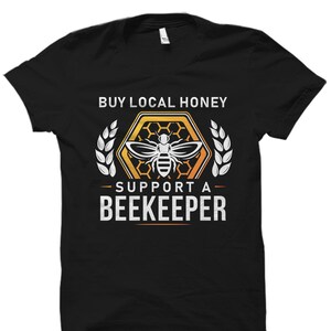 May include: Black t-shirt with a white graphic design of a bee in a honeycomb. The text "Buy Local Honey - Support a Beekeeper" is printed on the shirt.