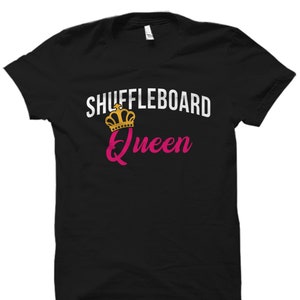 Puede incluir: Camiseta negra con texto blanco que dice "Shuffleboard Queen" y un gráfico de corona dorada.