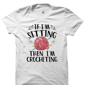 May include: White t-shirt with black text that reads "If I'm sitting then I'm crocheting". A pink ball of yarn with needles is pictured above the text.