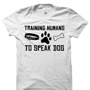 May include: White t-shirt with black text that reads "Training Humans to Speak Dog" with a black collar, paw print, and bone graphic.