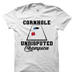 May include: White t-shirt with black text that reads "Cornhole Undisputed Champion". A black and white illustration of a cornhole board with a red and blue bean bag is in the center of the shirt.