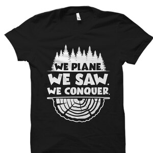 以下が含まれることがあります： 白いグラフィックデザインが施された黒いTシャツ。森のシルエットと「We Plane. We Saw. We Conquer.」というテキストが描かれています。