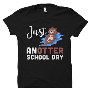 May include: Black t-shirt with a cartoon otter floating on its back in blue water. The text "Just another school day" is printed in white on the shirt.