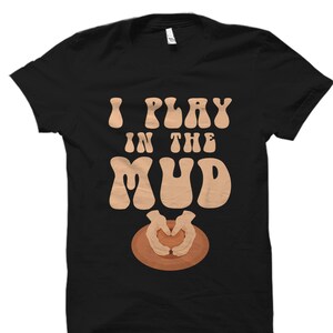 May include: Black t-shirt with the text "I Play in the Mud" in a retro font. The text is in brown and the shirt features a brown clay pot with two hands forming a heart shape around it.