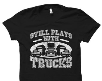 Può includere: Maglietta nera con una grafica bianca che dice "Still Plays With Trucks" e raffigura tre camion.