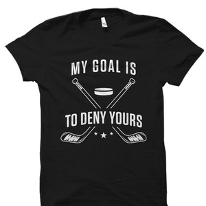 Może przedstawiać: Czarny T-shirt z białym nadrukiem przedstawiającym dwa skrzyżowane kije hokejowe nad krążkiem. Na koszulce widnieje napis "My goal is to deny yours".