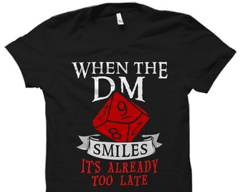 Peut inclure: T-shirt noir avec du texte blanc qui dit "When the DM smiles it's already too late". Un dé rouge et blanc est présenté dans le design.