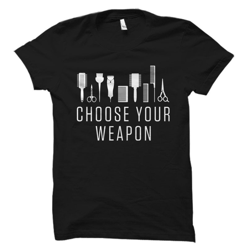 May include: Black t-shirt with a white graphic of various hairdressing tools, including scissors, combs, and clippers. The text "Choose Your Weapon" is printed below the tools.