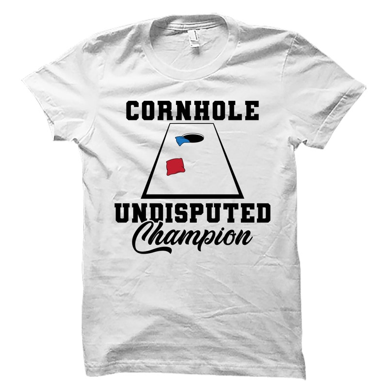 May include: White t-shirt with black text that reads "Cornhole Undisputed Champion". A black and white illustration of a cornhole board with a red and blue bean bag is in the center of the shirt.