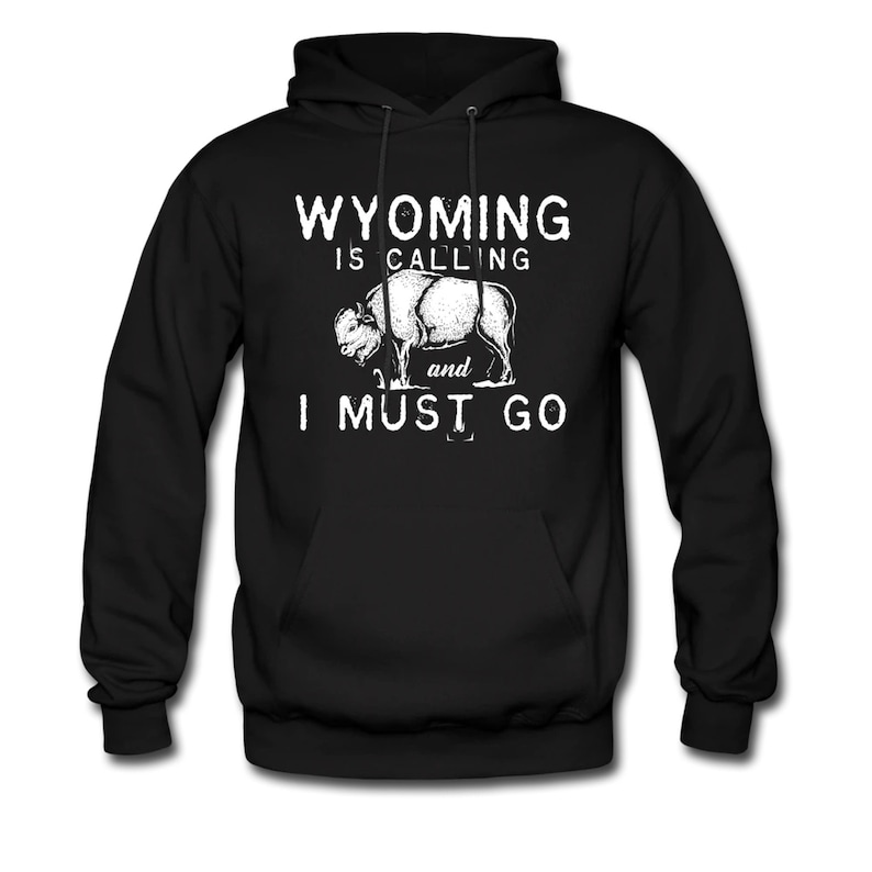 May include: Black hoodie with a white graphic. The graphic features a bison and the text "WYOMING IS CALLING and I MUST GO" in a distressed font. The hoodie has a drawstring hood and a front pocket.