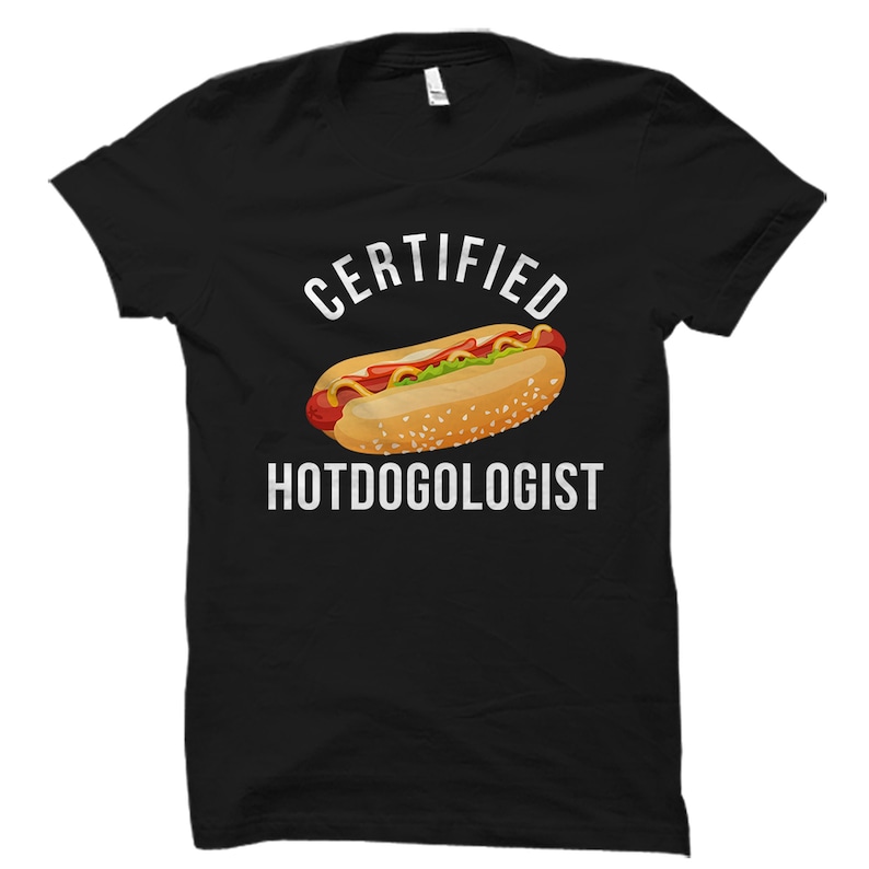 May include: Black t-shirt with white text that reads "CERTIFIED HOTDOGOLOGIST". A cartoon hot dog with mustard, ketchup, and relish is above the text.