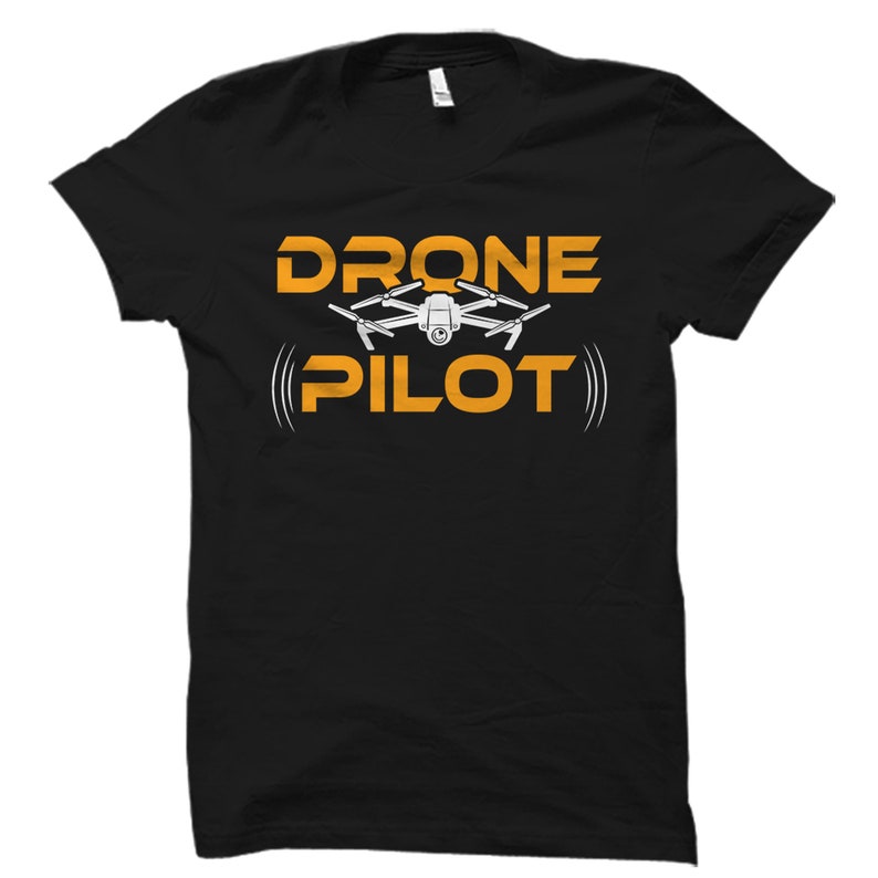 Puede incluir: Camiseta negra con el texto "DRONE PILOT" en letras naranjas. Un dron blanco con una c&aacute;mara est&aacute; por encima del texto.