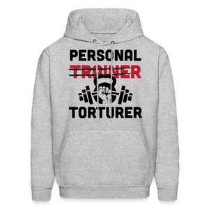 May include: A gray hooded sweatshirt with the text "PERSONAL TRAINER" crossed out and " TORTURER" written below it. A black and white illustration of a weightlifter holding a kettlebell is in between the text.