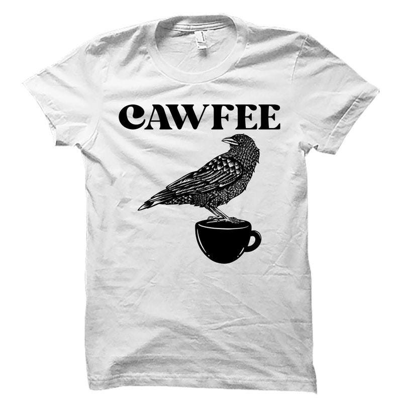 Pode incluir: Camiseta branca com uma ilustra&ccedil;&atilde;o preta de um corvo empoleirado em uma x&iacute;cara de caf&eacute;. O texto "CAWFFEE" est&aacute; impresso acima do corvo.