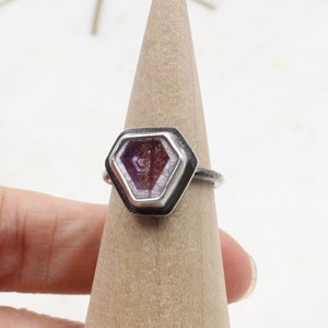 May include: A silver ring with a geometric, faceted, purple gemstone set in a black bezel. The ring is displayed on a wooden ring holder.