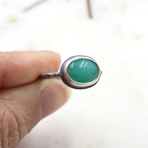 Op de afbeelding: Een zilveren ring met een groene ovale edelsteen in een bezel setting. De band van de ring is getextureerd.