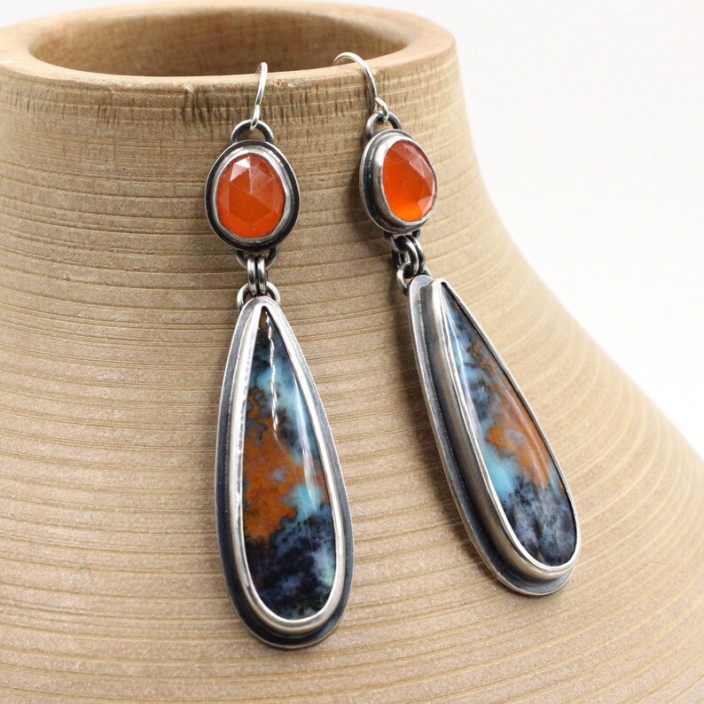 May include: A pair of silver dangle earrings with teardrop-shaped blue and orange agate stones and smaller orange carnelian stones at the top.
