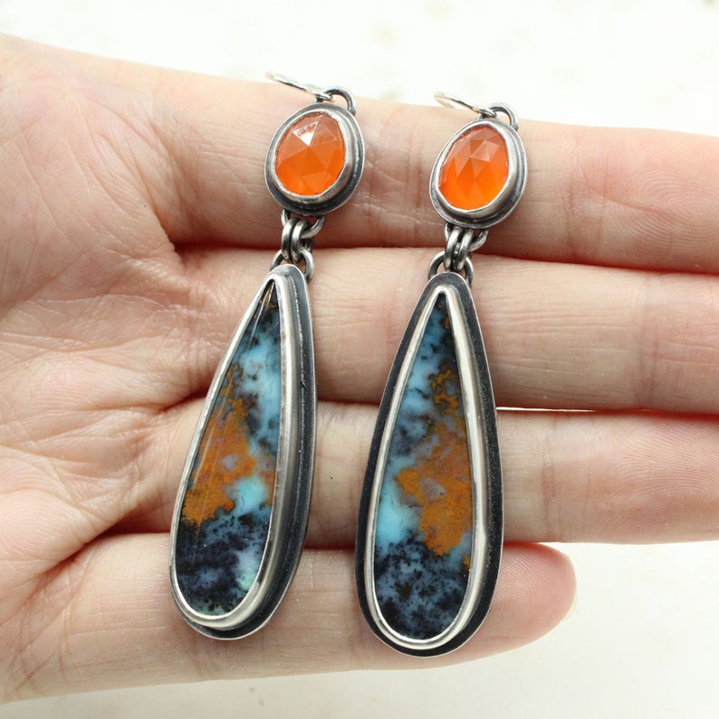 May include: A pair of silver dangle earrings with teardrop-shaped orange and blue agate stones and orange carnelian accents.