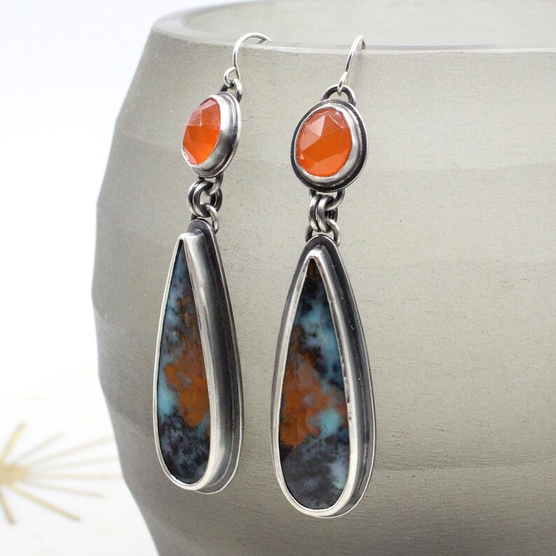 May include: A pair of silver teardrop earrings with orange gemstone accents. The earrings feature a teardrop-shaped stone with a brown, blue, and white pattern.