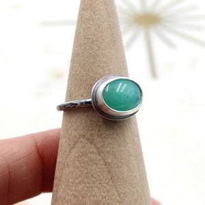Op de afbeelding: Een zilveren ring met een groene ovale edelsteen in een bezel-setting. De band van de ring is getextureerd.