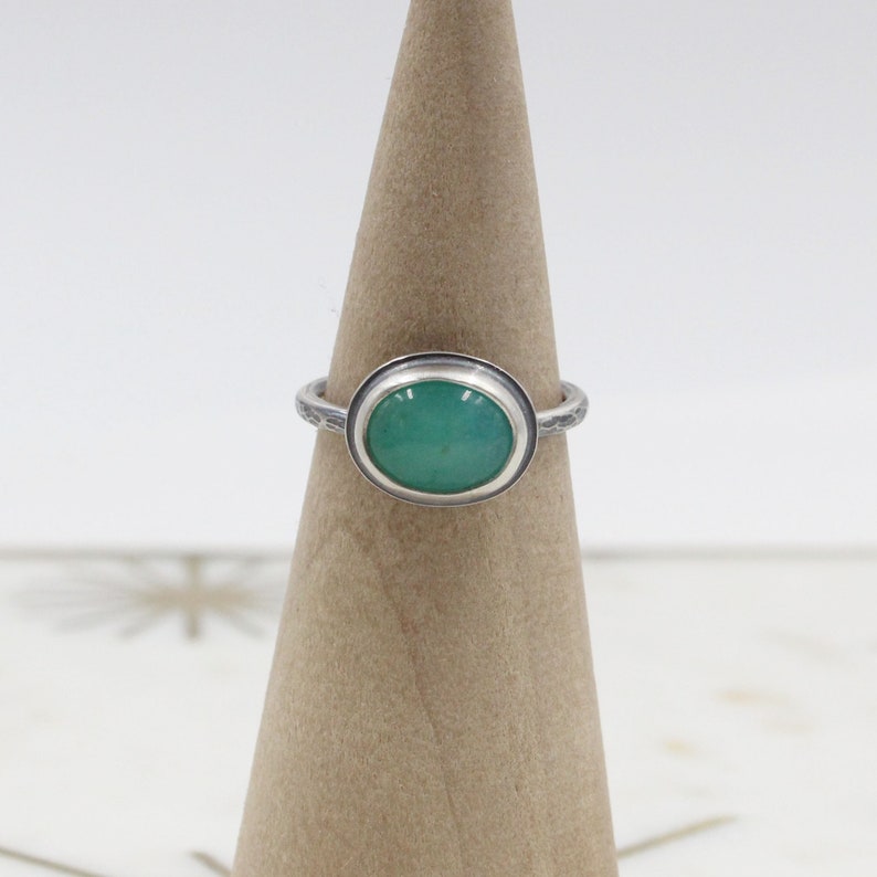 Op de afbeelding: Een zilveren ring met een groene ovale edelsteen in een bezel-setting. De ringband is getextureerd.