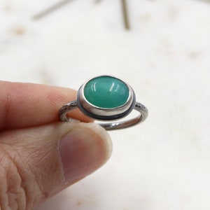 Op de afbeelding: Een zilveren ring met een groene ovale edelsteen in een bezel-setting. De band van de ring heeft een getextureerde afwerking.