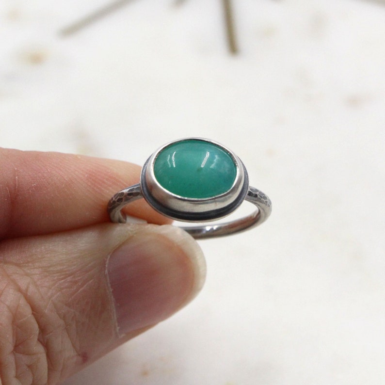 Op de afbeelding: Een zilveren ring met een groene ovale edelsteen in een bezel-setting. De band van de ring heeft een getextureerde afwerking.