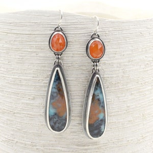 May include: A pair of silver teardrop earrings with a blue and brown agate stone and a small orange carnelian gemstone at the top.