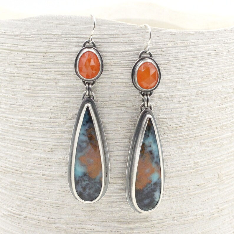 May include: A pair of silver teardrop earrings with a blue and brown agate stone and a small orange carnelian gemstone at the top.