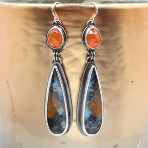 May include: A pair of silver teardrop earrings with orange gemstone accents. The teardrop stones are a mix of blue, brown, and white.