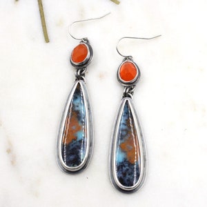 May include: A pair of silver dangle earrings with teardrop-shaped stones. The stones are a mix of blue, orange, and brown with a silver border. The top of each earring features a smaller round orange stone.