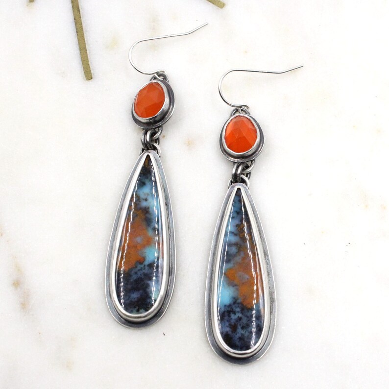 May include: A pair of silver dangle earrings with teardrop-shaped stones. The stones are a mix of blue, orange, and brown with a silver border. The top of each earring features a smaller round orange stone.