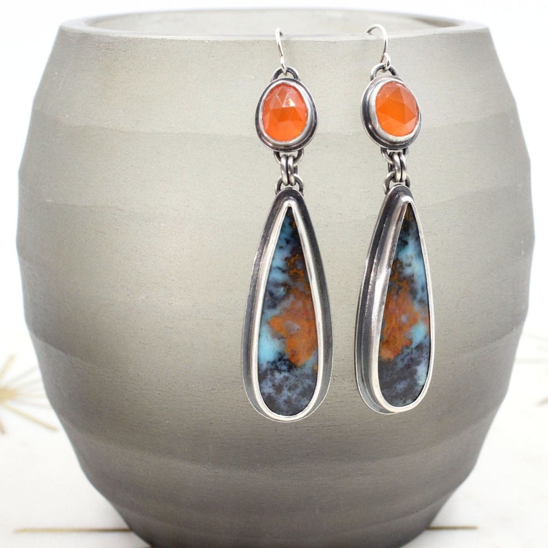 May include: A pair of silver teardrop earrings with a blue and orange agate stone and a smaller orange gemstone at the top.