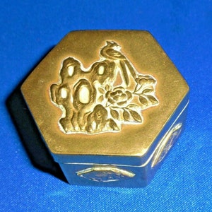 May include: A small hexagonal brass trinket box with an embossed design of a bird perched on a branch with a flower. The box has a gold finish and is decorated with a white enamel design.