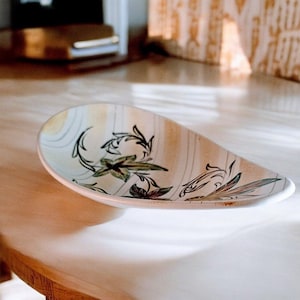 May include: A white ceramic serving dish with a leaf design in shades of green and brown. The dish is oval shaped and has a slightly raised rim.