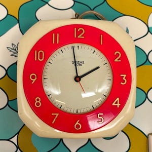 May include: A square-shaped Smiths Electric wall clock with rounded corners, featuring a cream-coloured outer frame and a bright red circular dial. The clock face is cream with gold-coloured numerals from 1 to 12, black hour and minute hands, and a thin red second hand.