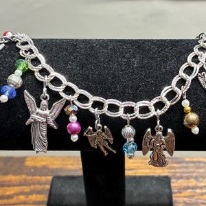 May include: Silver-toned charm bracelet featuring angel pendants and colorful beads. The bracelet has a chain link design and is displayed on a black surface. The charms include various angel designs and decorative beads in shades of blue, pink, and green.