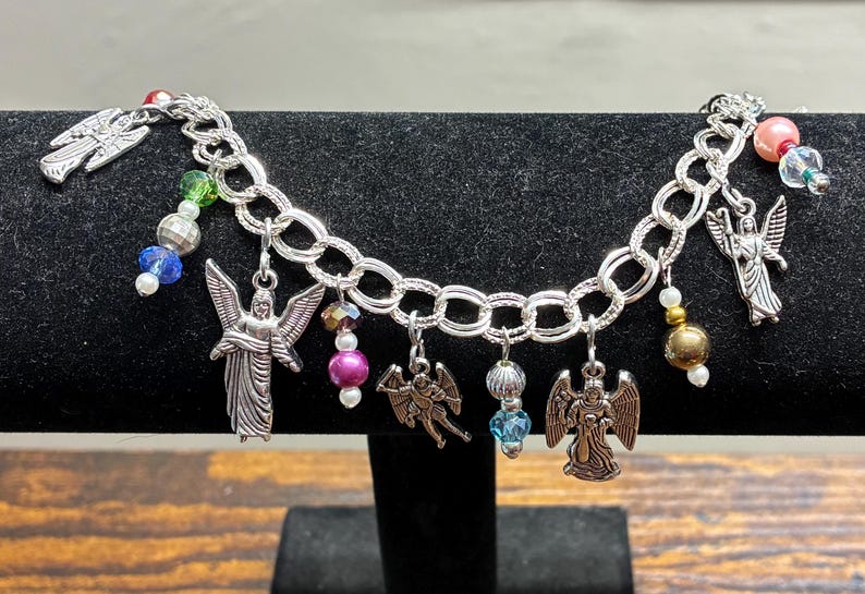 May include: Silver-toned charm bracelet featuring angel pendants and colorful beads. The bracelet has a chain link design and is displayed on a black surface. The charms include various angel designs and decorative beads in shades of blue, pink, and green.