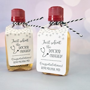 May include: Two small glass bottles with red caps and white labels. The labels have black text that reads "Just what the doctor ordered!" and "Congratulations! [Name] M.D."