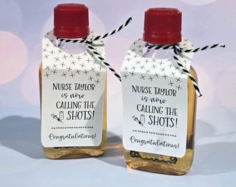 Könnte beinhalten: Zwei kleine, durchsichtige Glasflaschen mit roten Kappen und weißen Etiketten. Die Etiketten haben schwarzen Text, der "Nurse Taylor is now calling the shots! Congratulations!" lautet.